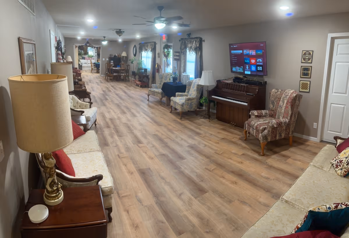 A long, well-lit common area in a residential care center with wooden flooring, multiple upholstered chairs and sofas, a piano with a flat-screen TV mounted above it, and several tables and lamps. The space has a cozy and inviting atmosphere with neutral-colored walls and decorative curtains on the windows.