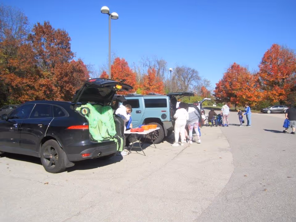 A parking lot scene with several cars parked with their trunks open. People are gathered around the cars, some standing and some sitting. There is a table set up with items on it next to one of the cars. The background shows trees with autumn-colored leaves under a clear blue sky.