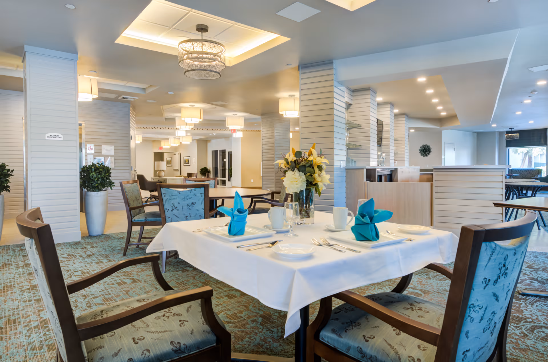 Bright, modern dining room with a table set for four, a floral centerpiece, and upholstered chairs in a senior living facility.