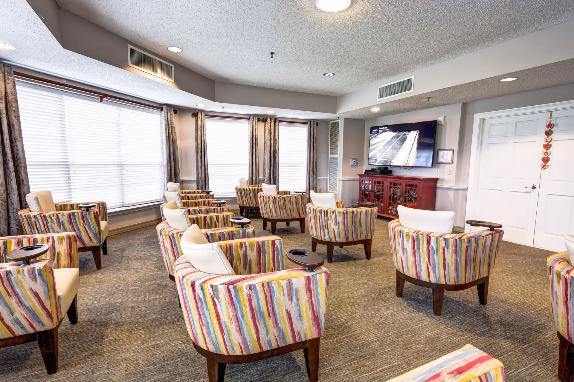 A bright common area with multiple colorful striped armchairs arranged in rows facing a wall-mounted flat screen TV. The room has large windows with blinds and curtains, beige carpet, and a red cabinet beneath the TV. A white door with a hanging welcome decoration is visible on the right side.