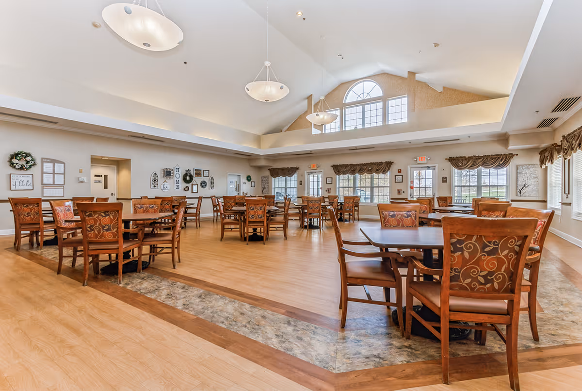 Spacious dining room with wooden tables and chairs arranged neatly on a wood and tile floor. The room has high vaulted ceilings with large hanging light fixtures and large windows with valance curtains letting in natural light. Walls are decorated with framed pictures and wreaths.