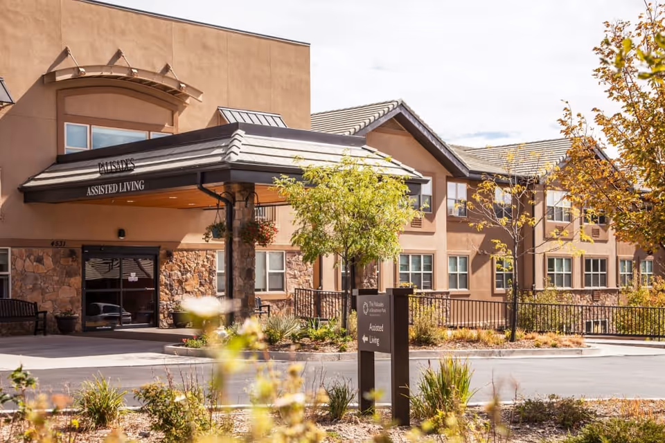Front entrance of a beige assisted living building with a covered porte-cochere, landscaped grounds, and a sign.