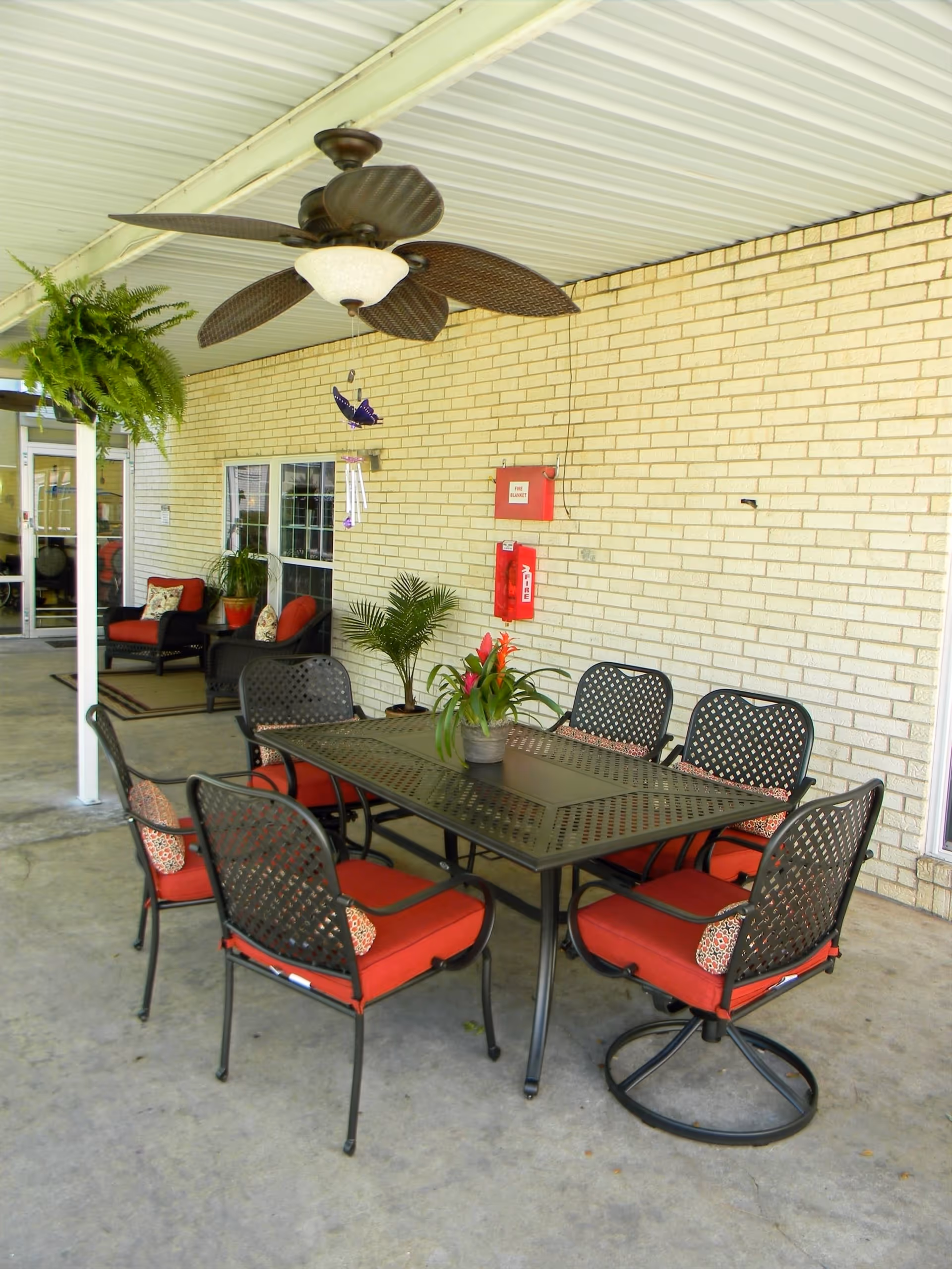 Covered outdoor patio area with a black metal table and six matching chairs with red cushions. There is a ceiling fan above the table, potted plants, and a brick wall with a fire alarm and fire extinguisher mounted on it. In the background, there are additional seating areas with cushioned chairs and a hanging fern plant.