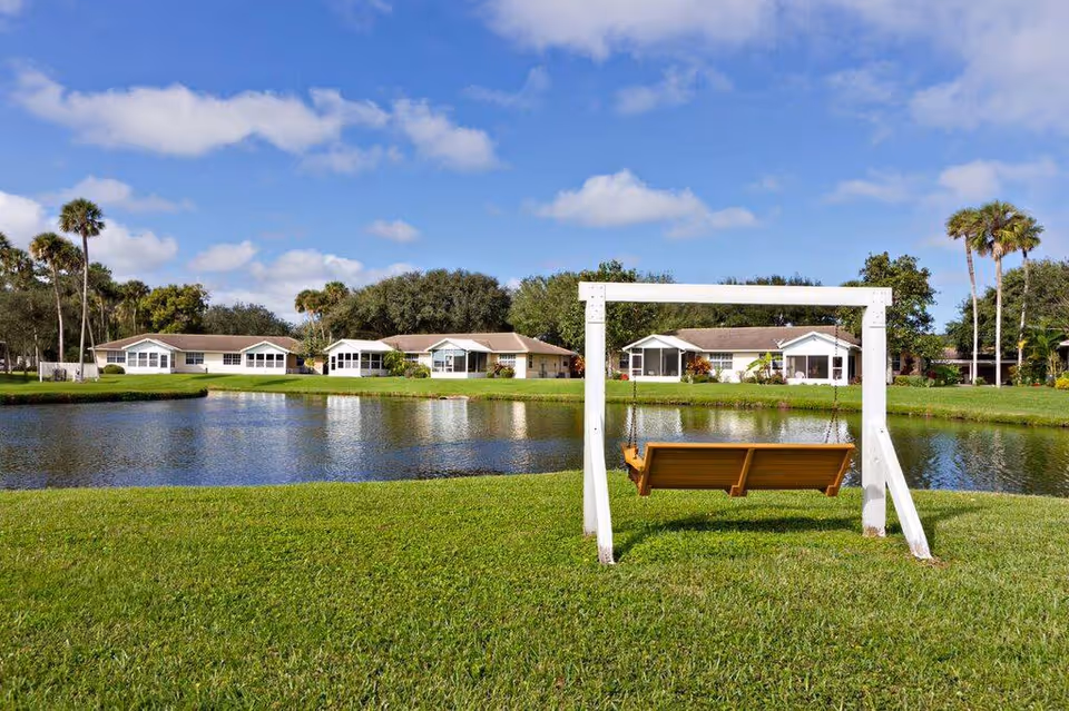A peaceful outdoor scene at Hidden Lakes Senior Living Community featuring a wooden swing on a white frame facing a small lake with several single-story houses and palm trees in the background under a partly cloudy blue sky.