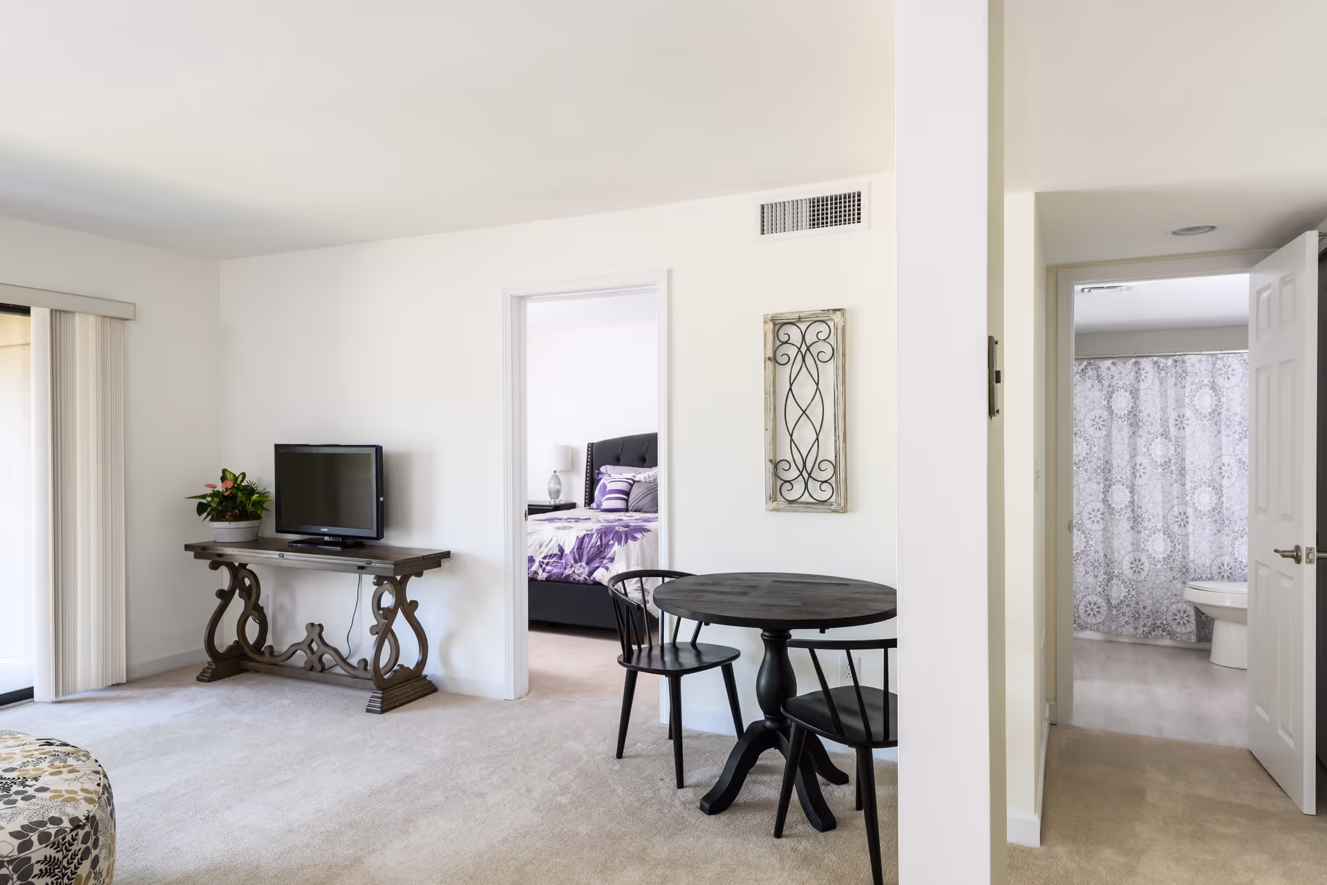 Interior view of a senior living facility showing a living room area with a small round table and two chairs, a TV on a decorative wooden console table, a bedroom with a bed covered in purple bedding visible through an open door, and a bathroom with a shower curtain and toilet visible through another open door.