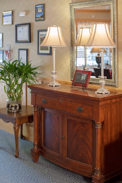 A wooden sideboard with two decorative table lamps and a framed photo on top, positioned in front of a large ornate mirror. To the left, there is a small wooden table with a potted plant. The wall behind features framed certificates and artwork.