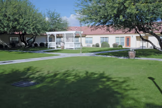 A well-maintained outdoor courtyard area with green grass, a white gazebo, paved walkways, trees, and a single-story building with a red roof and multiple windows in the background.