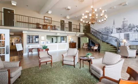Spacious senior living facility lobby with comfortable armchairs and small tables arranged on a green patterned carpet. A large chandelier hangs from the ceiling, and a staircase with green carpet leads to an upper balcony area. The reception desk is visible in the background with a large mural of a town skyline on the wall behind the staircase.
