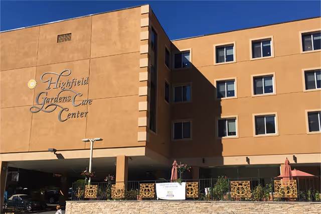 Exterior view of a multi-story building with tan stucco walls and multiple windows under a clear blue sky. The building has a sign that reads 'Highfield Gardens Care Center' on the left side. There is a covered area at the ground level with some parked cars and a patio area with umbrellas and plants in front.