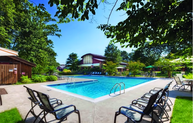 Outdoor swimming pool surrounded by lounge chairs and trees, with a building visible in the background under a clear blue sky.