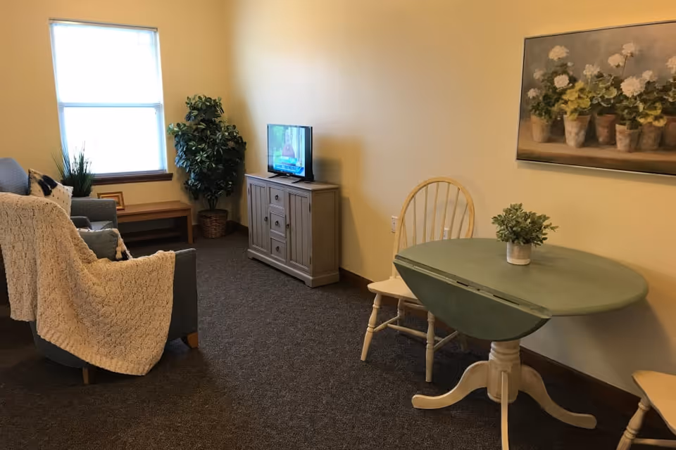 A cozy living room area with a blue armchair draped with a cream-colored knitted blanket, a small wooden bench under a window, a potted plant in the corner, a wooden TV stand with a flat-screen TV, a round green table with two wooden chairs, and a floral painting on the wall.