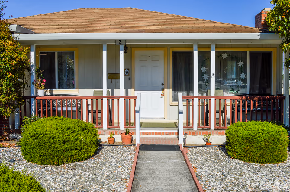 Front exterior view of a single-story residential building with a small porch, white door, two windows with curtains, potted plants, and neatly trimmed bushes on either side of a concrete walkway leading to the entrance under a clear blue sky.