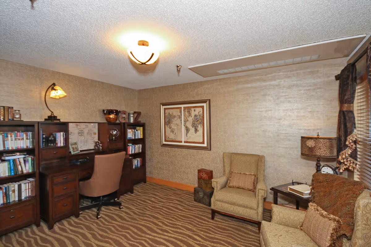 A cozy interior room featuring a wooden desk with a brown swivel chair, bookshelves filled with books, a beige armchair with a decorative pillow, a side table with a lamp and clock, and a framed world map on the wall. The room has patterned carpet and textured beige walls with a ceiling light fixture.