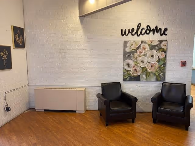 A cozy corner of a room with two black leather armchairs placed on a wooden floor against a white brick wall. Above the chairs, there is a decorative sign that says 'welcome' and a painting of white and pink flowers in a vase. On the left side of the wall, there are two small framed artworks with botanical designs. A beige radiator cover is positioned below the artworks.