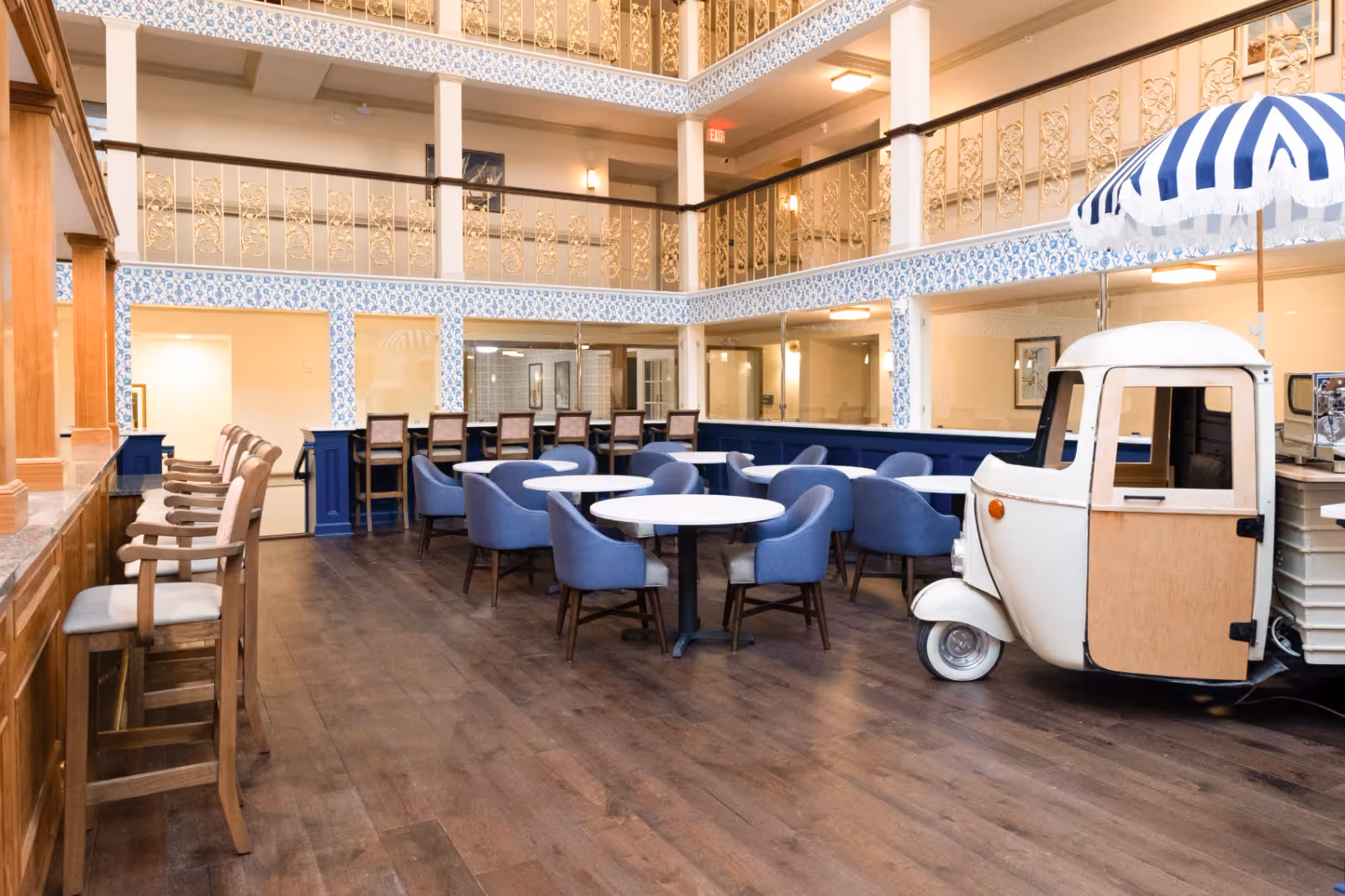 Interior view of a spacious dining area with multiple round tables surrounded by blue upholstered chairs. There are also wooden bar stools lined up along a counter on the left side. The room features decorative railings on the upper level and blue and white patterned wallpaper accents. A small white vehicle with a blue and white striped umbrella is parked on the right side of the room.
