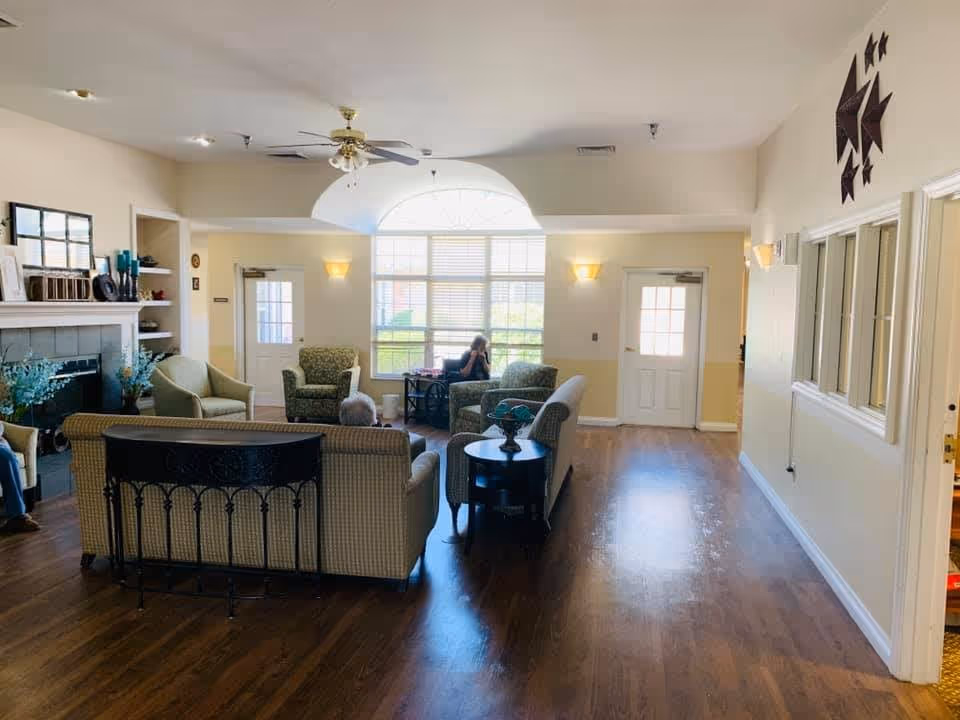 A bright and spacious living room area in a senior living facility with several cushioned armchairs and sofas arranged around a fireplace. There is a large window letting in natural light, a ceiling fan, wooden flooring, and a person sitting near the window. The walls are painted light yellow and white, and there are decorative items on the fireplace mantel and a star-shaped wall decoration.
