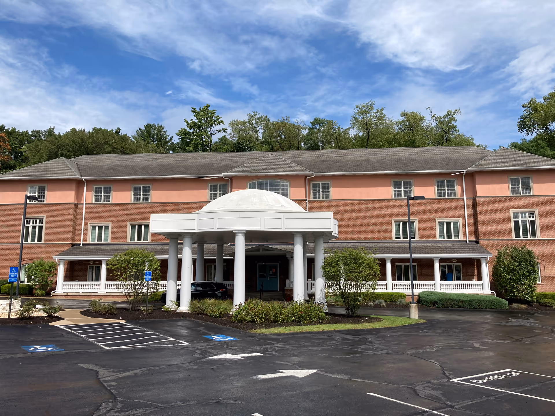 Front entrance of a three-story brick senior living building with a white domed portico, driveway, and parking area.