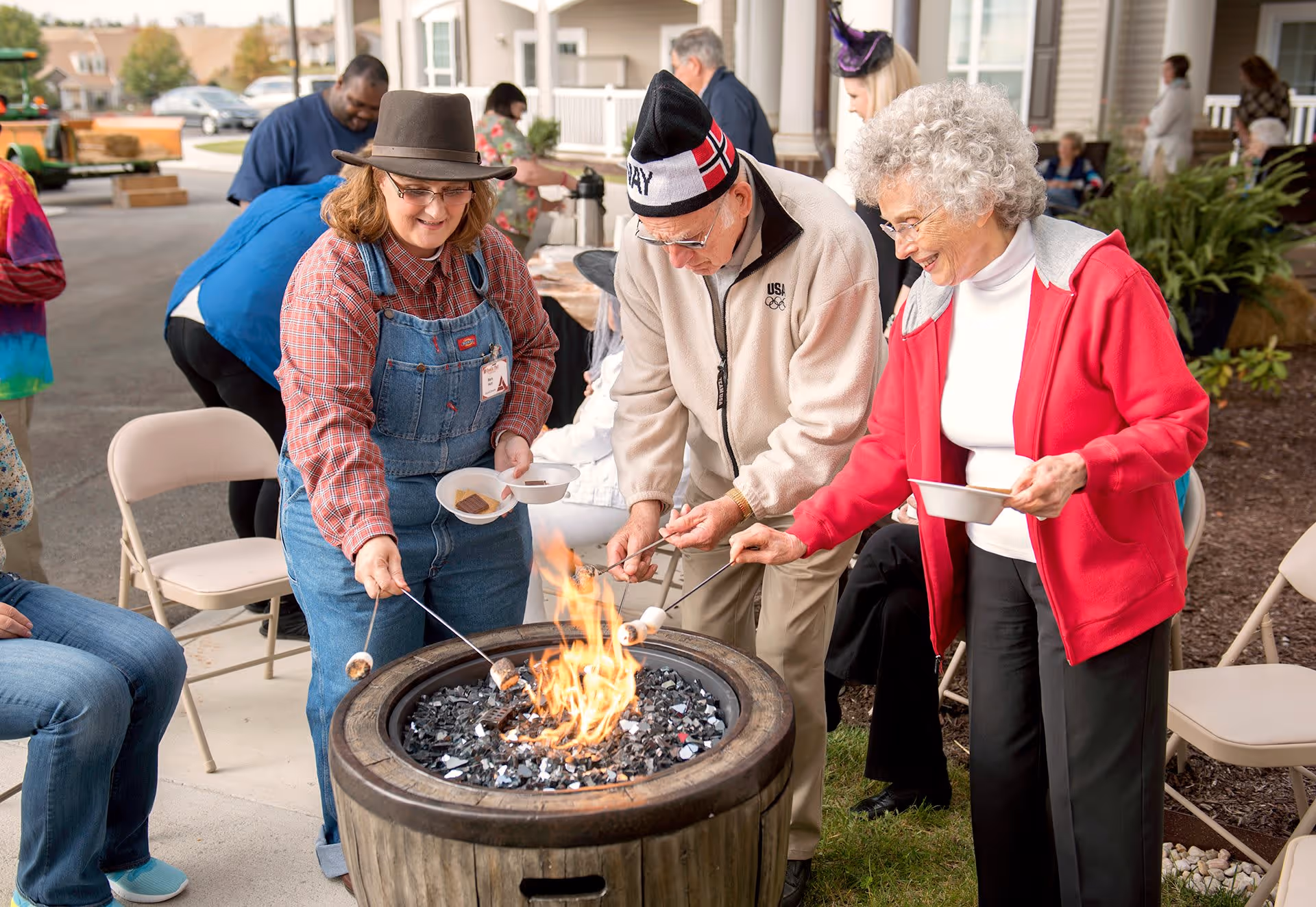 Three elderly people roasting marshmallows over an outdoor fire pit. One woman is wearing a red jacket and white turtleneck, an elderly man is wearing a beige jacket and a winter hat, and another woman is wearing a plaid shirt with denim overalls and a brown hat. They are smiling and enjoying the activity, with chairs and other people visible in the background near a building.