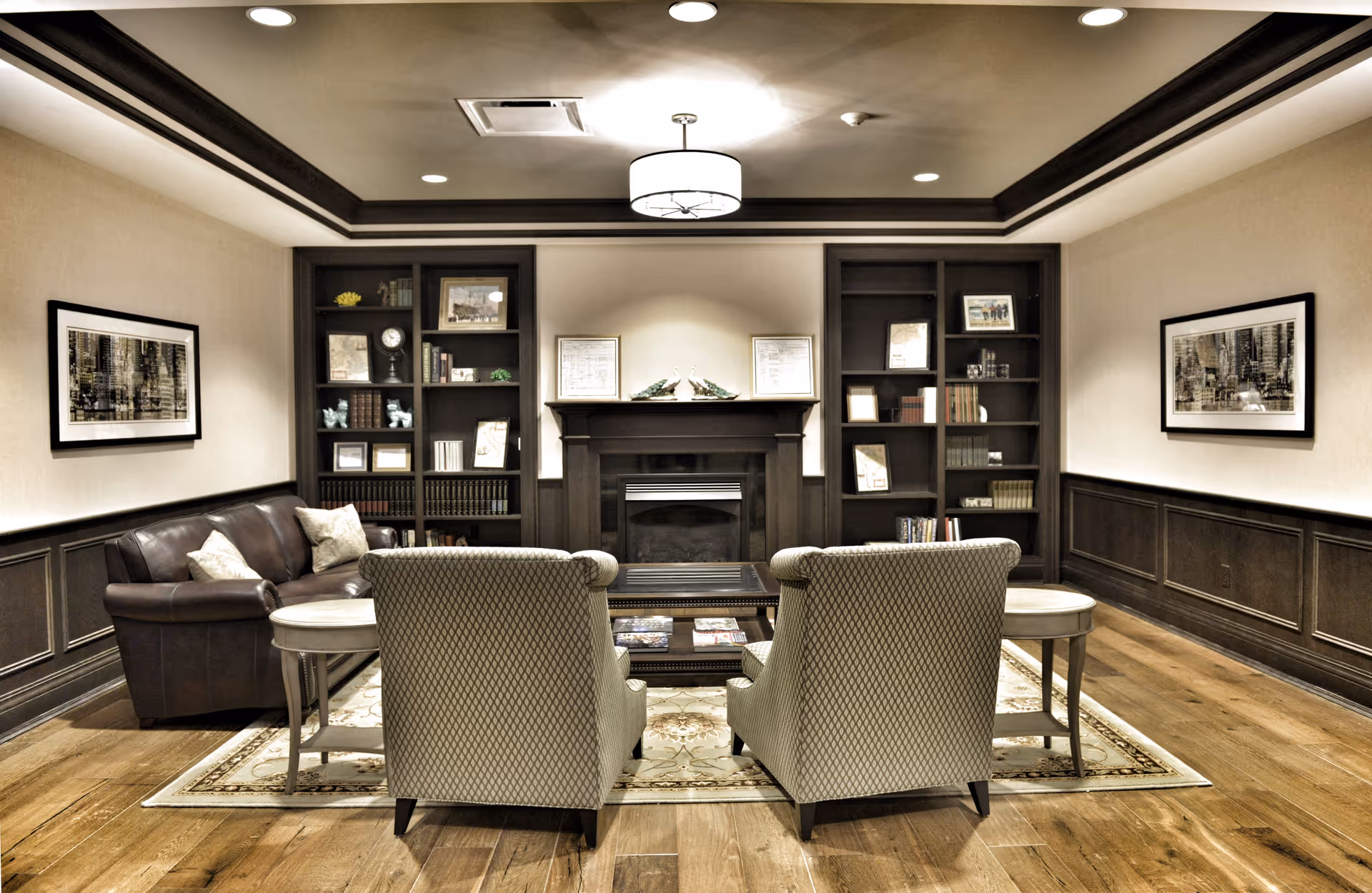 A cozy living room with two patterned armchairs and a brown leather sofa arranged around a wooden coffee table on a decorative rug. The room features built-in dark wood bookshelves flanking a fireplace, framed artwork on the walls, and recessed lighting in the ceiling.