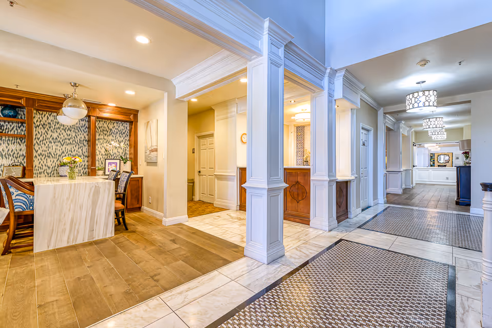 Bright senior living facility interior showing a lobby/reception area with seating, a dining nook, and tiled floors.