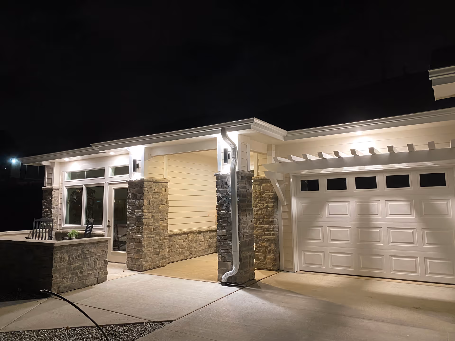 Night view of the exterior entrance of a facility with stone pillars, a white garage door, and a small patio area with chairs and a table. The building is illuminated by outdoor lights.