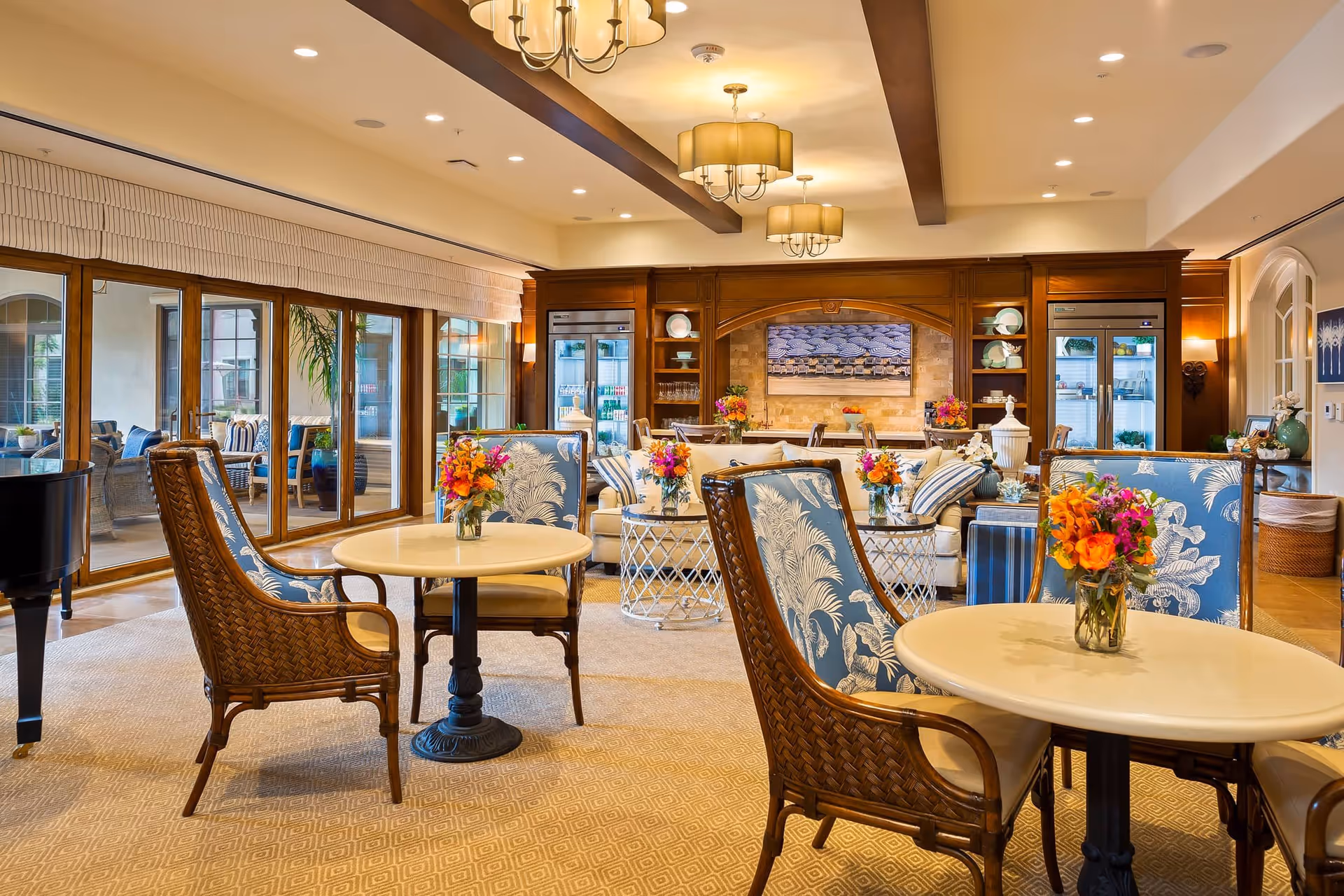 A cozy and elegant common area with round tables and wicker chairs featuring blue and white patterned cushions. Each table has a small vase with colorful flowers. The room has warm lighting with ceiling chandeliers and large windows with wooden frames that open to an outdoor seating area. In the background, there is a wooden built-in cabinet with glass doors, shelves holding dishes, and a large framed artwork on the wall.