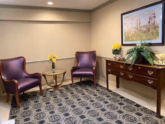 A small sitting area with two purple armchairs and a round glass-top side table with a vase of yellow flowers. There is a wooden console table with drawers against the wall, decorated with a potted plant, a yellow flower arrangement, and a small horse figurine. A framed floral painting hangs above the console table. The floor is covered with a patterned carpet and the walls are beige with a chair rail.