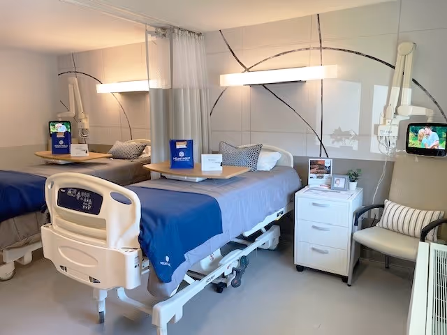 A clean and well-lit hospital or rehabilitation room with two beds, each equipped with blue bedding and pillows. Each bed has a bedside table with informational materials and a mounted adjustable light above. There is a white three-drawer cabinet between the beds and a comfortable chair with a striped pillow next to it. The walls have a modern design with curved black lines on a light background.