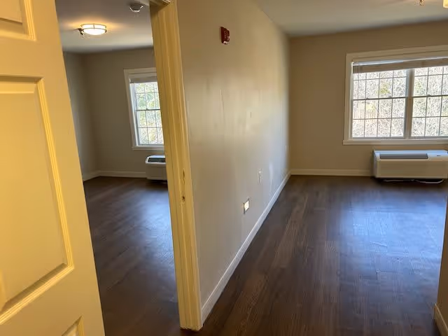 View of an empty interior space in Wellington Manor Knoxville showing two rooms with wooden flooring, beige walls, and windows with white blinds. One room is partially visible through an open door on the left, and the other room is fully visible on the right with a heating or air conditioning unit below the window.