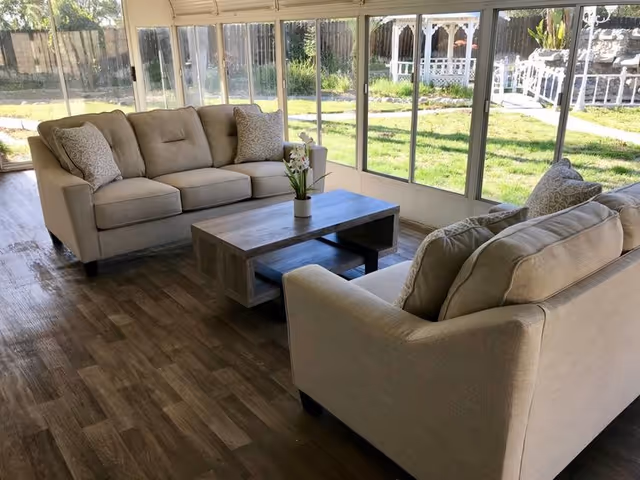Sunlit enclosed sunroom with two beige sofas facing a wooden coffee table and large windows overlooking a grassy yard.