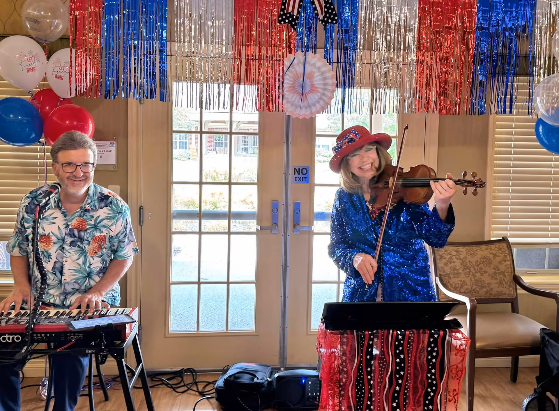 Two musicians — a keyboard player and a violinist in a sparkly blue outfit and red hat — perform in a decorated indoor common room with patriotic streamers and balloons.