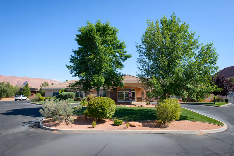 Exterior view of a single-story assisted living facility building with a landscaped front yard featuring green trees, bushes, and a small lawn. The building has a covered entrance with an American flag displayed. The sky is clear and blue, and there are mountains visible in the background.