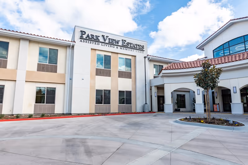 Exterior front view of Park View Estates Assisted Living & Memory Care building with a clear sky and a small landscaped area with a tree near the entrance.