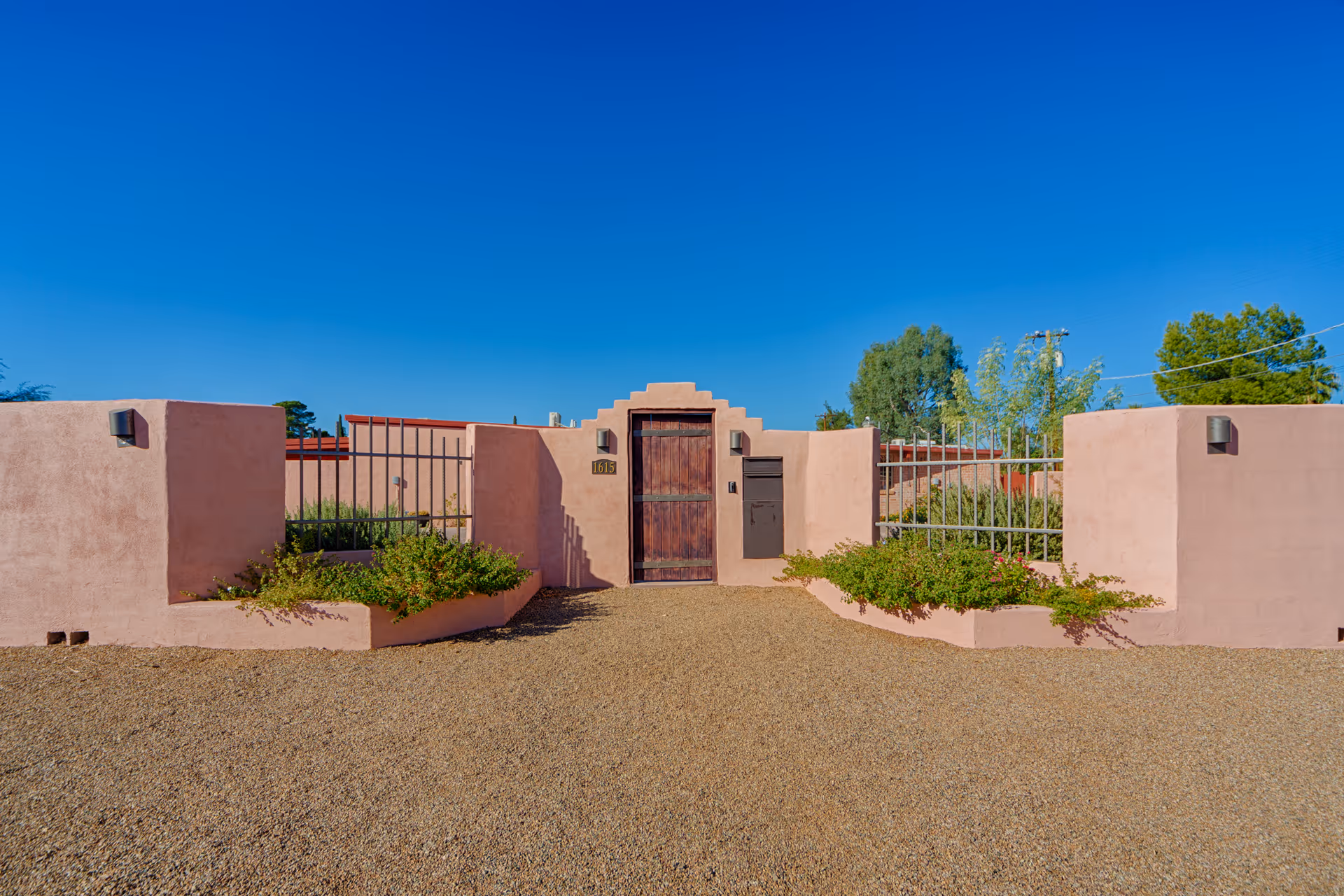 Exterior view of a southwestern-style entrance gate with pink stucco walls, a wooden door, metal railings, and small garden beds with green plants on either side under a clear blue sky.