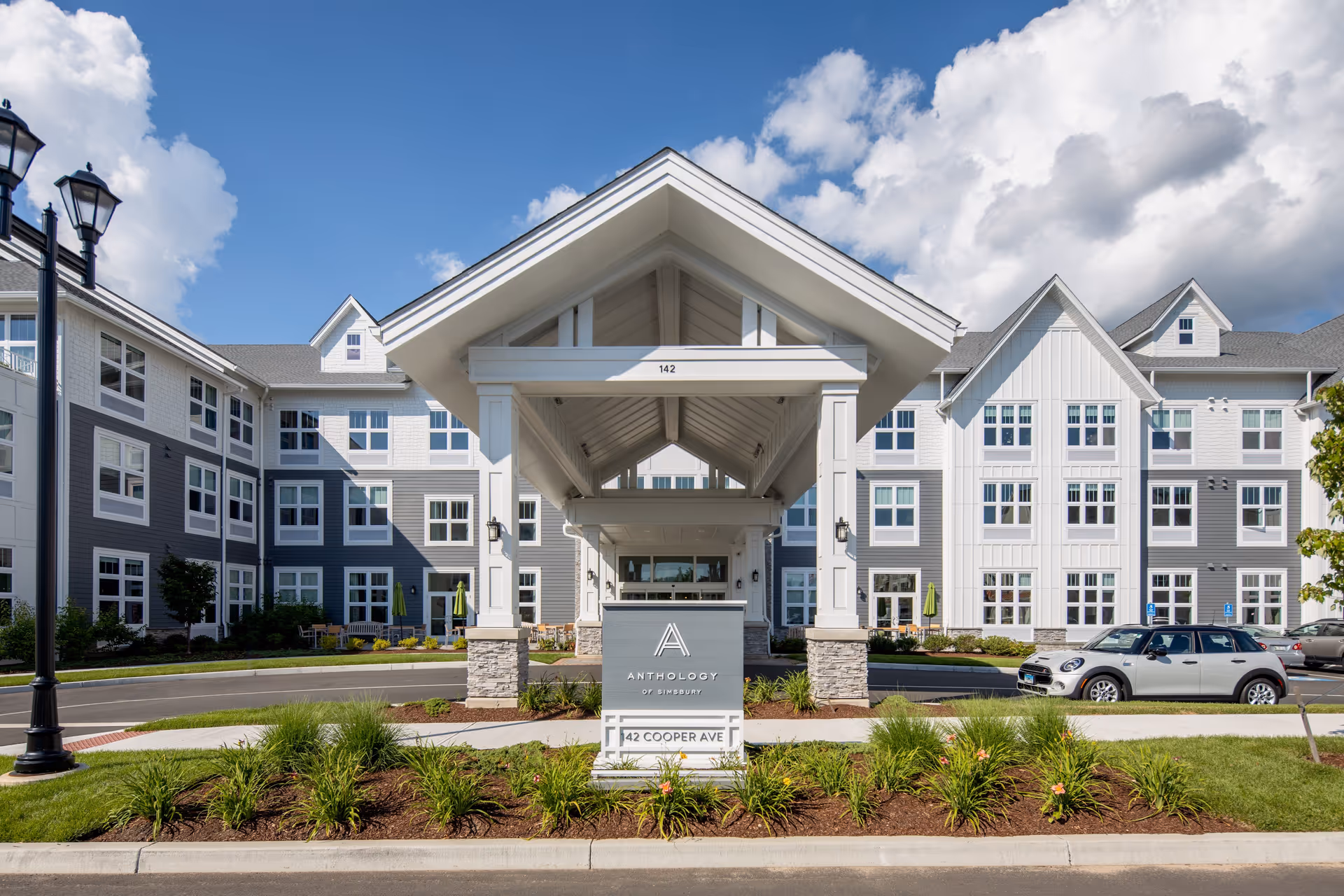 Front exterior view of a senior living facility named Anthology of Simsbury, featuring a covered entrance with white pillars and stone bases, surrounded by landscaped greenery and a driveway with parked cars under a partly cloudy sky.