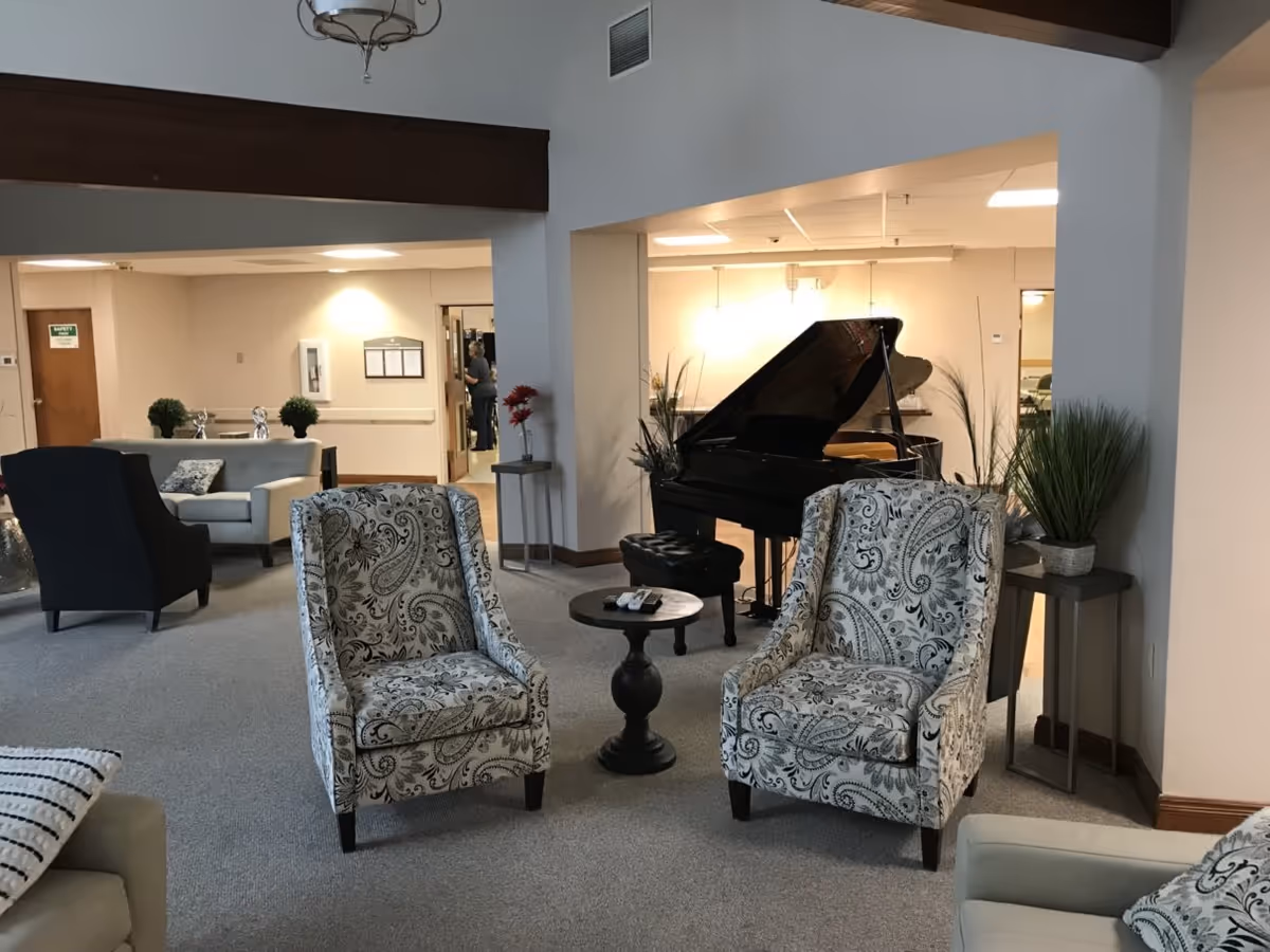 A cozy living room area in Good Shepherd Care Center featuring two patterned armchairs with a small round table between them, a black grand piano in the background, beige sofas, potted plants, and soft lighting.