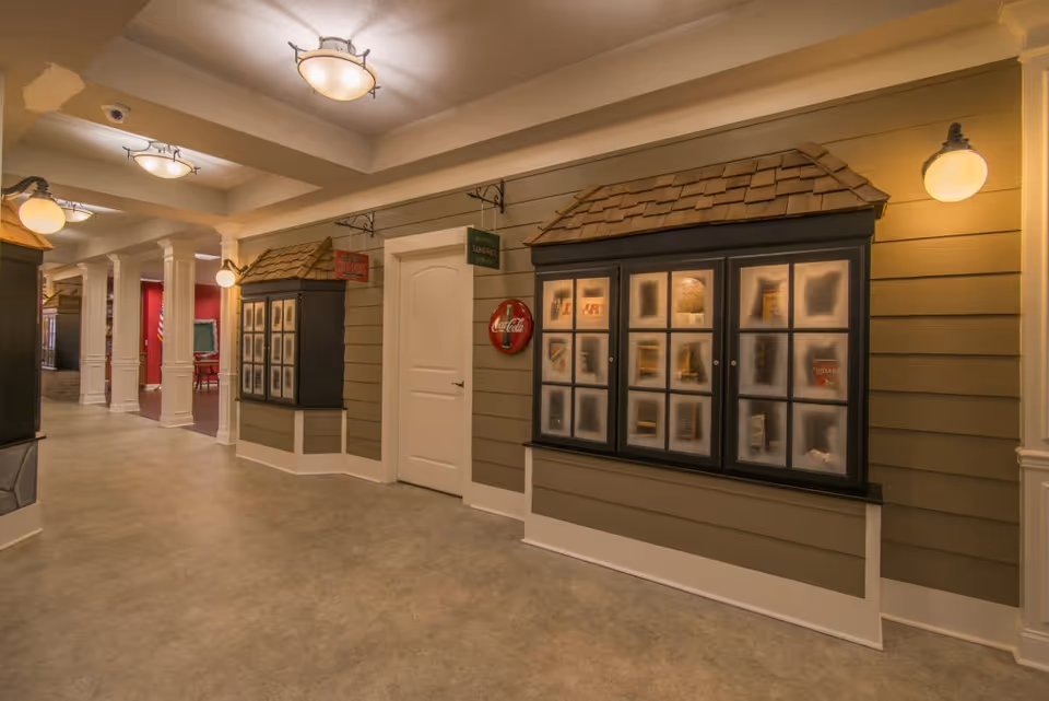 Well-lit interior hallway with storefront-style display windows, a white door, and decorative wall sconces.