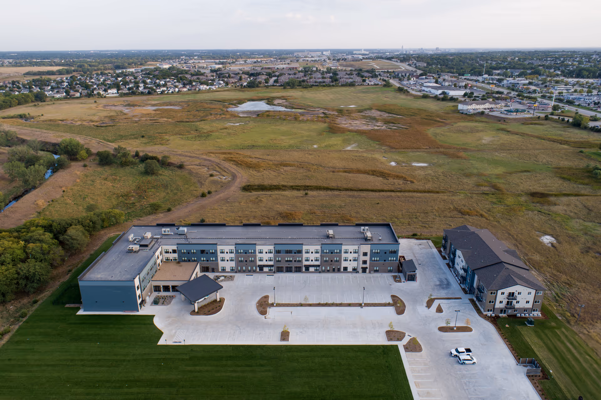 Aerial view of Salt Creek Senior Living facility showing a large, modern three-story building with a spacious parking lot in front, surrounded by green grass and open fields with a distant view of a suburban area.