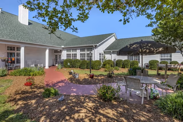 Outdoor courtyard area of Vickery Parke Assisted Living with a paved walkway, garden beds, bushes, and a seating area with a table and umbrella under a clear blue sky.