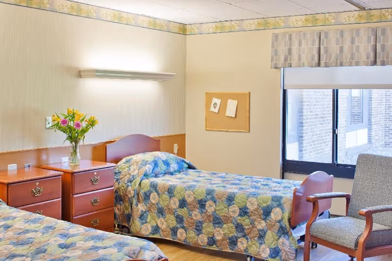 A bright twin bedroom with two beds, wooden nightstands, a window, and a chair in a senior living facility.