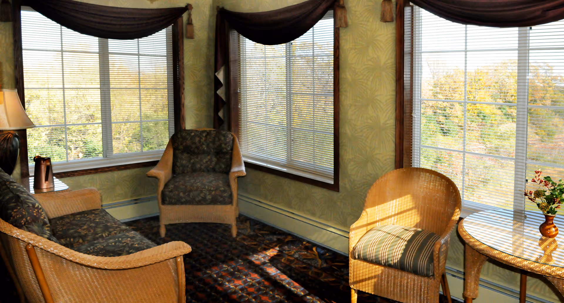 A cozy sitting area with wicker furniture including a sofa, two chairs, and a glass-top side table with a small vase of flowers. The room has large windows with blinds and dark valances, allowing natural light to fill the space and offering a view of trees outside.