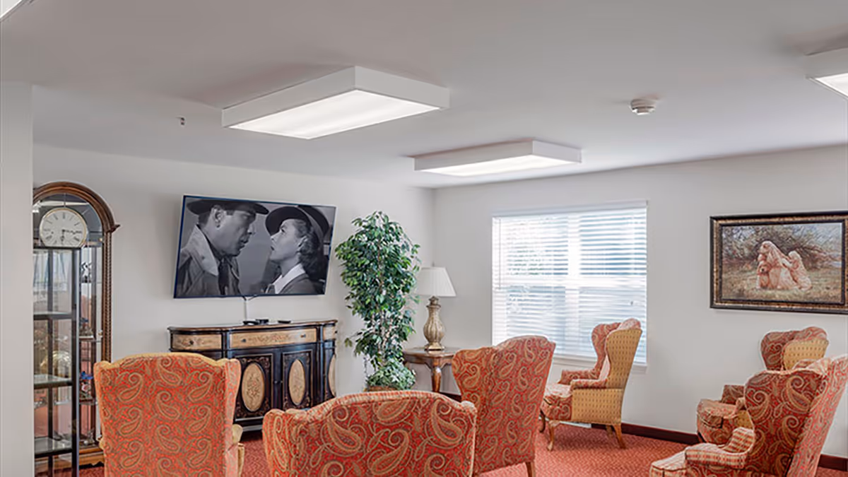 Well-lit communal sitting room with patterned armchairs, a wall-mounted TV, a grandfather clock, and framed art.
