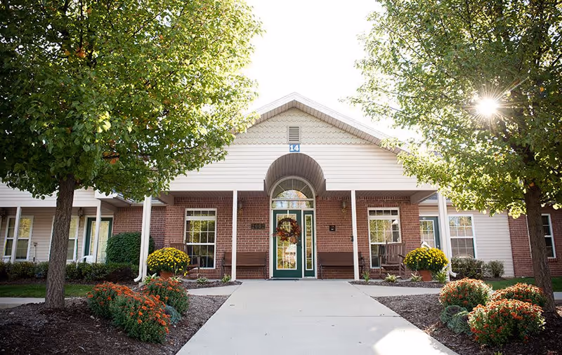 Front exterior view of a single-story assisted living facility building with a central entrance door decorated with a wreath. The building has a combination of brick and siding, with two large trees and landscaped flower beds on either side of the concrete walkway leading to the entrance.