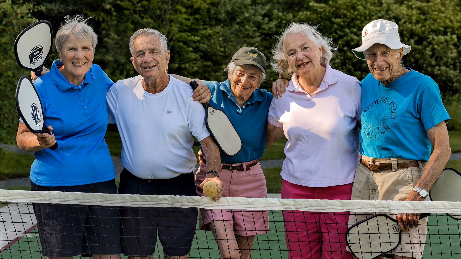 Five elderly adults standing together on a pickleball court outdoors, smiling and holding pickleball paddles, with a pickleball net in front of them and greenery in the background.