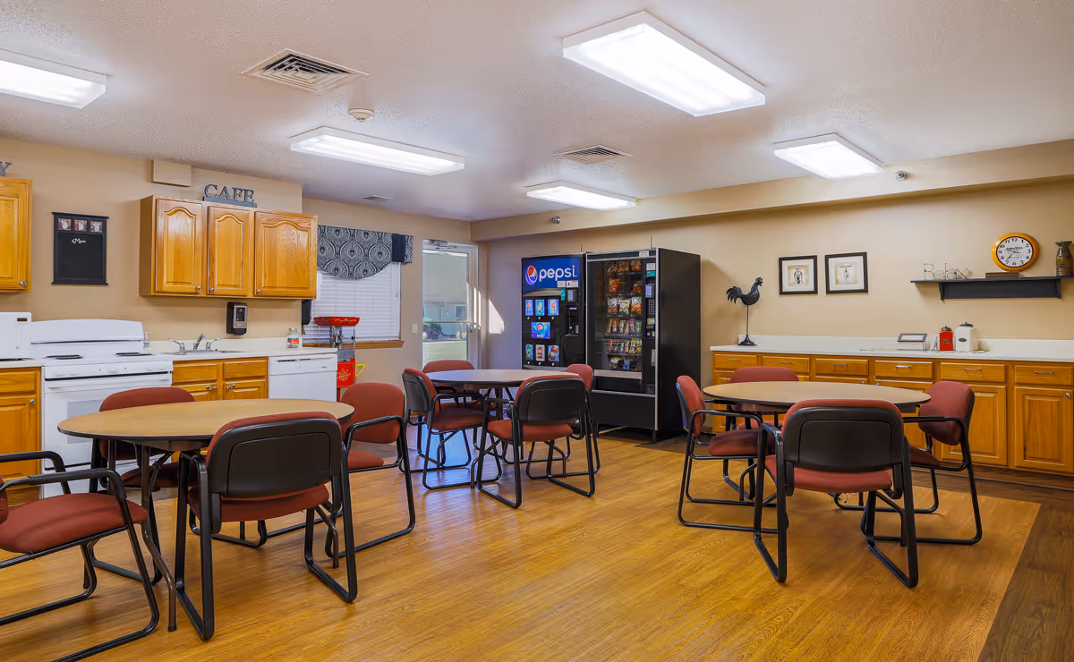 A bright communal dining area with round tables and red cushioned chairs. The room features wooden cabinets, a white stove, dishwasher, and microwave along one wall. There are vending machines for snacks and drinks near a window and door. The walls have simple decor including framed pictures, a clock, and a shelf with the word 'Relax'.