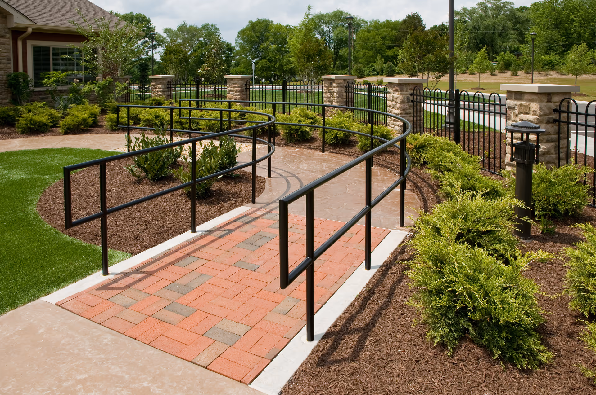 Outdoor garden area with a curved black metal handrail ramp leading to a building entrance. The pathway is paved with red and gray bricks, surrounded by mulch beds with green shrubs and small trees. Stone pillars and a black metal fence enclose the area, with a grassy lawn and trees in the background under a partly cloudy sky.