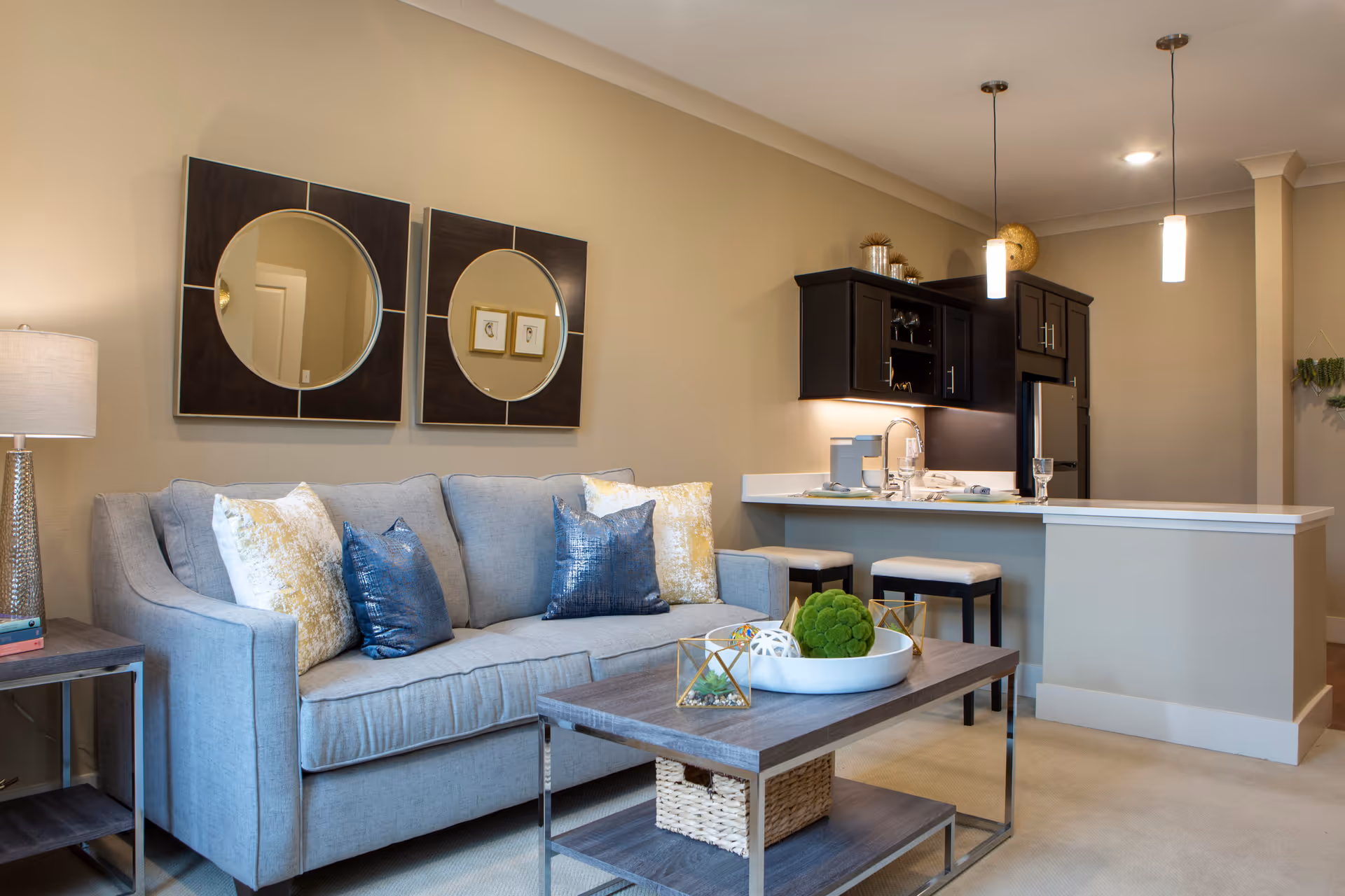 A modern living room area with a light gray sofa adorned with blue and yellow decorative pillows. Above the sofa are two square wall mirrors with circular reflective centers. In front of the sofa is a wooden coffee table with decorative items including a white bowl with green moss balls and geometric gold accents. To the right is a kitchen area with a white countertop, two bar stools, dark wood cabinets, a sink, and pendant lights hanging from the ceiling. A table lamp is visible on a side table to the left of the sofa.