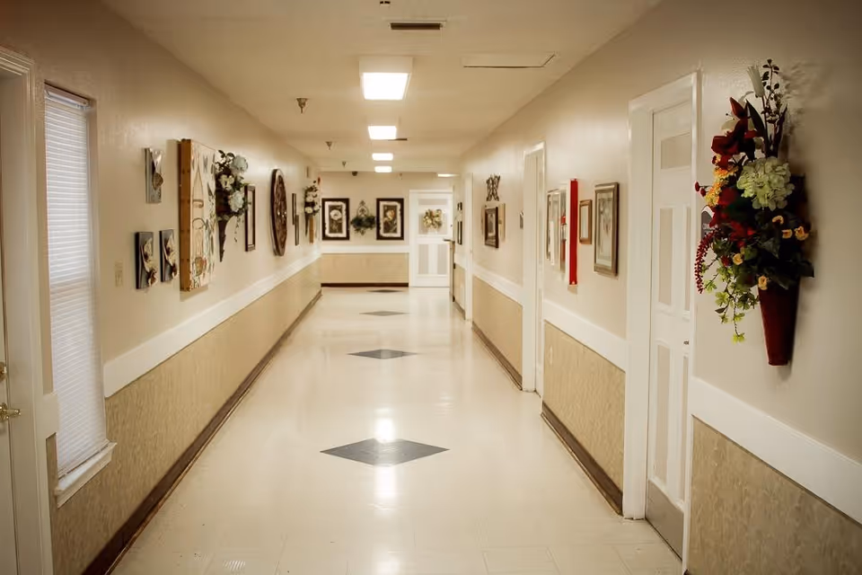 A long, well-lit hallway in a senior living facility with beige walls and floor tiles featuring diamond patterns. The walls are decorated with framed pictures and floral arrangements. Several closed doors line the hallway on the right side, and a window with blinds is on the left.