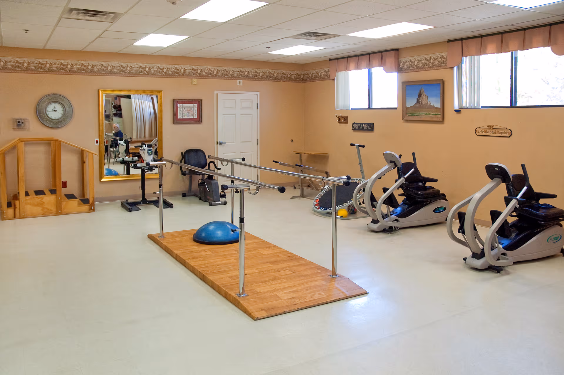 A rehabilitation or exercise room with parallel bars on a wooden platform, exercise bikes, a blue exercise ball, and a small wooden ramp. The room has beige walls with framed pictures and windows letting in natural light.