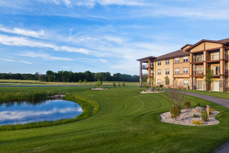 A large, three-story residential building with balconies overlooking a well-maintained green lawn and a small pond. The sky is blue with some clouds, and there are trees in the background.